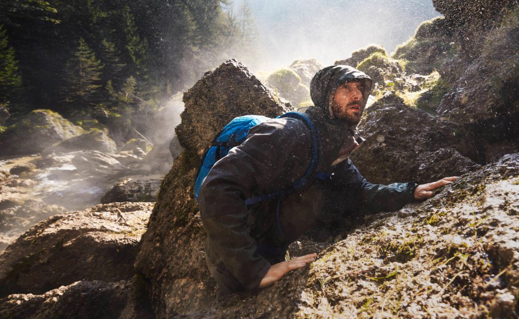 Wildniswandern 3 Wanderer mit schwarzer Regenjacke bei einer kurzen Klettertour an einem Wasserfall.