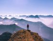 Eine Person auf dem Gipfel eines Berges. Vor der Person erstrecken sich spektakuläre Gebirgslandschaften.