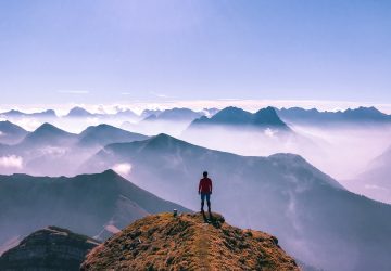 Eine Person auf dem Gipfel eines Berges. Vor der Person erstrecken sich spektakuläre Gebirgslandschaften.