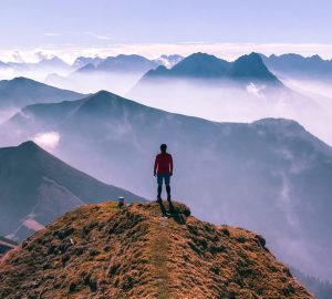 Eine Person auf dem Gipfel eines Berges. Vor der Person erstrecken sich spektakuläre Gebirgslandschaften.