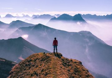 Eine Person auf dem Gipfel eines Berges. Vor der Person erstrecken sich spektakuläre Gebirgslandschaften.