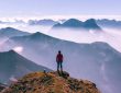 Eine Person auf dem Gipfel eines Berges. Vor der Person erstrecken sich spektakuläre Gebirgslandschaften.