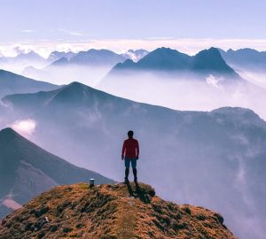Bergsicherheit 2 Eine Person auf dem Gipfel eines Berges. Vor der Person erstrecken sich spektakuläre Gebirgslandschaften.