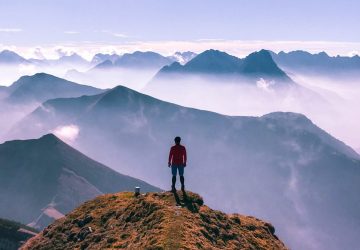 Eine Person auf dem Gipfel eines Berges. Vor der Person erstrecken sich spektakuläre Gebirgslandschaften.